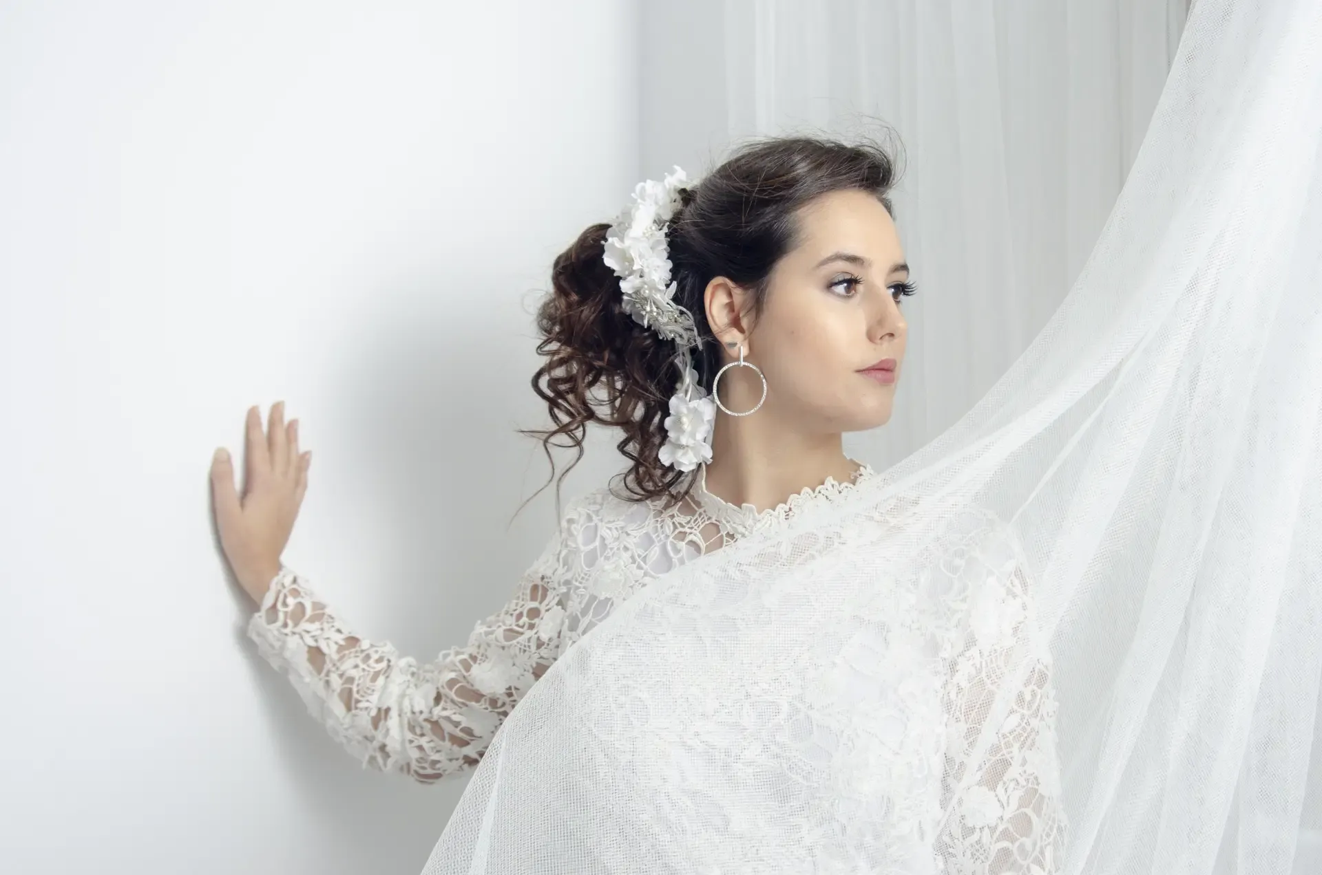 Editorial bridal photograph of a model in a white lace gown with veil, shot against soft white drapery