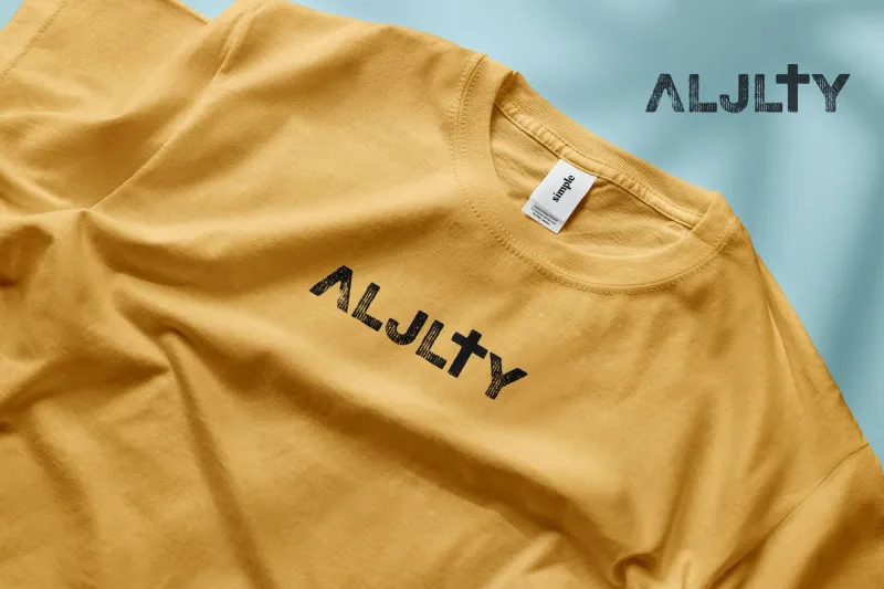 Close-up of a mustard gold t-shirt with the ALJLTY brand logo screen-printed in distressed black ink featuring a cross integrated into the letterforms, photographed at an angle on a light blue background